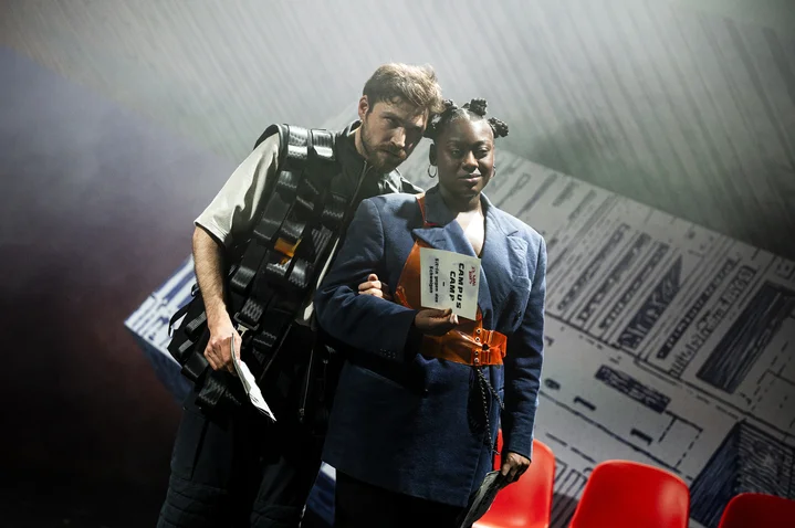 Two individuals on stage in dramatic lighting. One holds a book, with a backdrop of documents behind them.