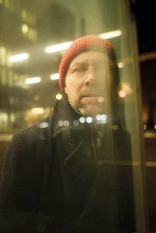 Ein Mann steht hinter einer Glasscheibe. Er trägt eine rote Mütze und dunkle Kleidung. Lichter spiegeln sich im Glas.