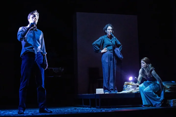 A theater scene with three actors on stage under blue lighting. Two are standing, one is seated. Dramatic atmosphere.