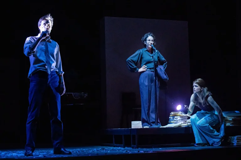 Eine Theaterszene mit drei Akteuren auf der Bühne, in blauer Beleuchtung. Zwei stehen, eine Person sitzt. Dramatische Atmosphäre.