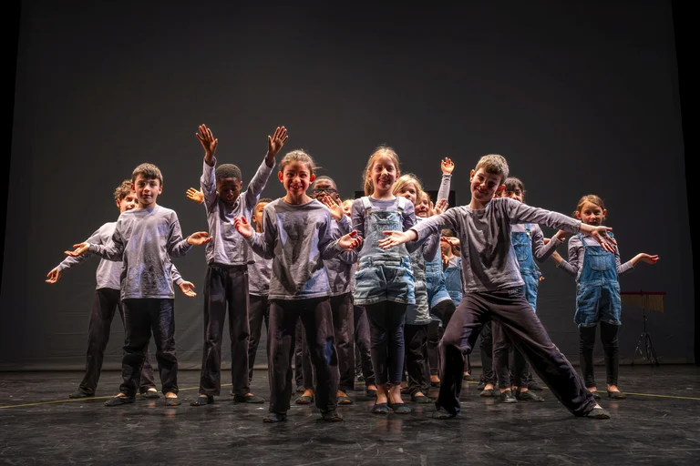 Eine Gruppe von Tänzern auf einer Bühne führt eine energetische Choreografie auf. Sie tragen lässige Kleidung und strahlen Freude aus.