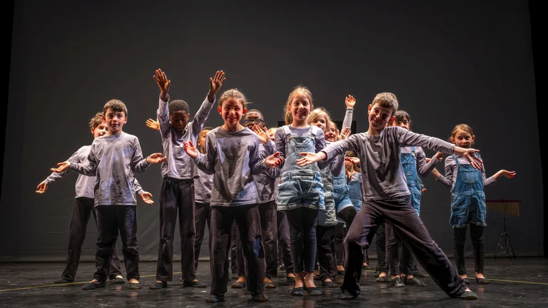 Eine Gruppe von Tänzern auf einer Bühne führt eine energetische Choreografie auf. Sie tragen lässige Kleidung und strahlen Freude aus.