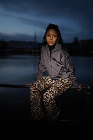 Person in Leopardenhose und grauer Jacke sitzt am Wasser bei Dämmerung, mit dunklem Himmel im Hintergrund.