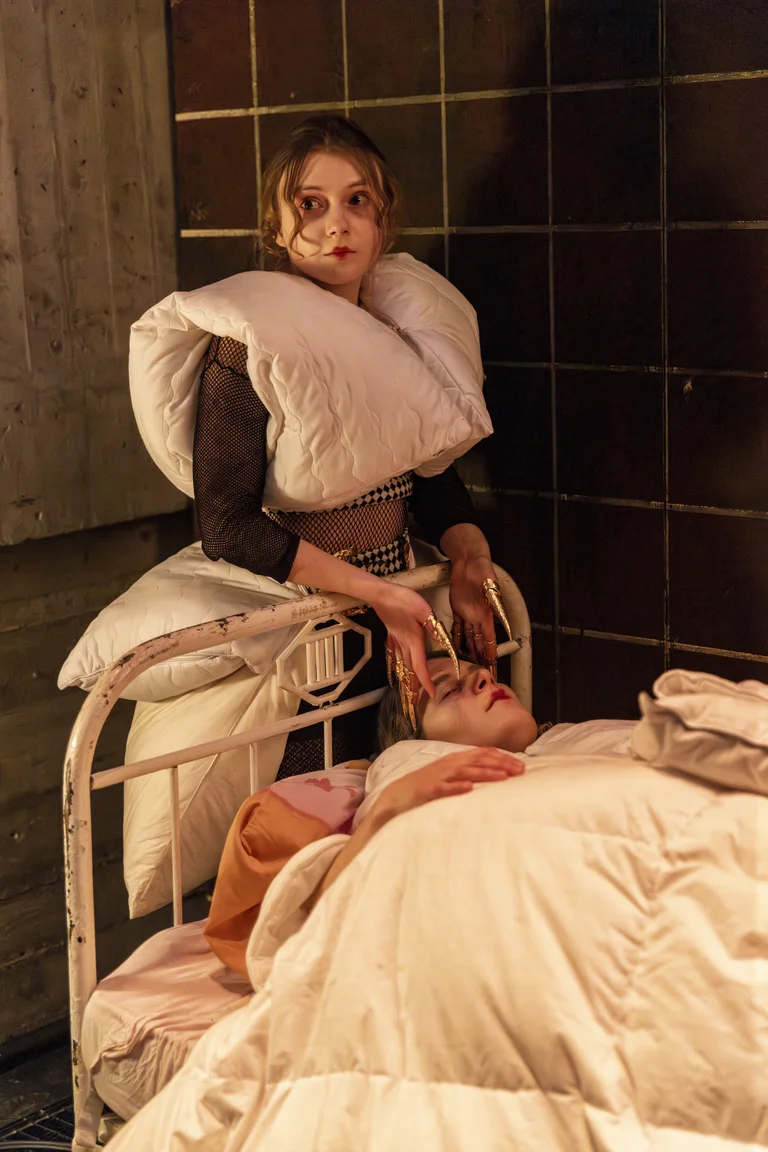 Eine Frau in Kissen gekleidet steht neben einem alten Bett und berührt den Kopf einer Person, die im Bett liegt.