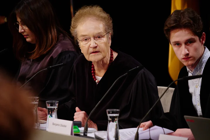 Eine Frau mittleren Alters spricht in einem formellen Rahmen, flankiert von zwei jüngeren Personen. Sie tragen Anzug und Mikrofon.