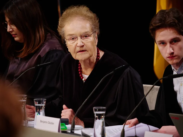 Eine Frau mittleren Alters spricht in einem formellen Rahmen, flankiert von zwei jüngeren Personen. Sie tragen Anzug und Mikrofon.