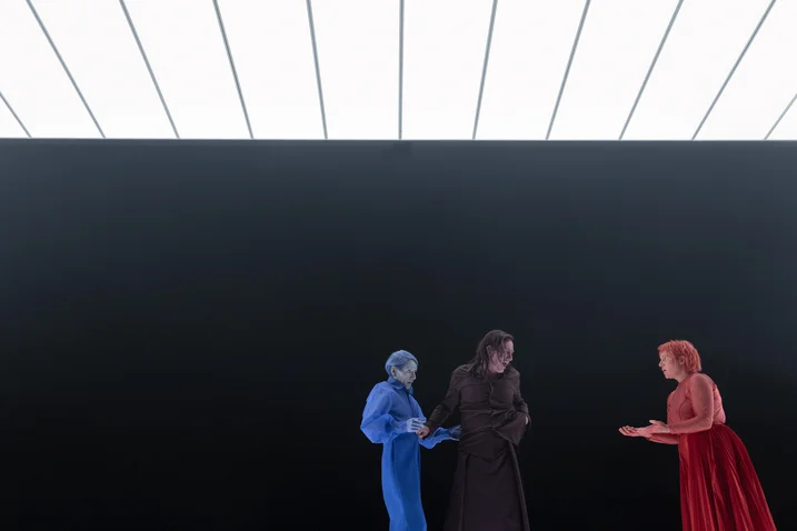 Three individuals in colorful costumes stand against a dark background. One is in blue, one in black, and one in red.