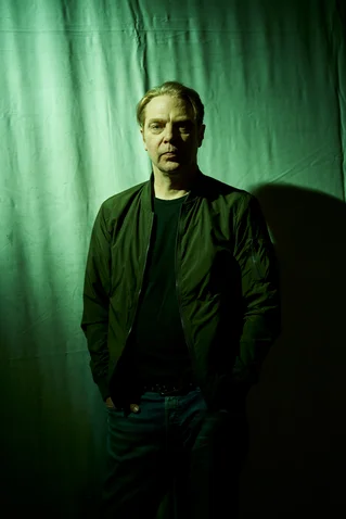 Ein Mann steht vor einem grün beleuchteten Hintergrund. Er trägt eine dunkle Jacke und eine Jeans.