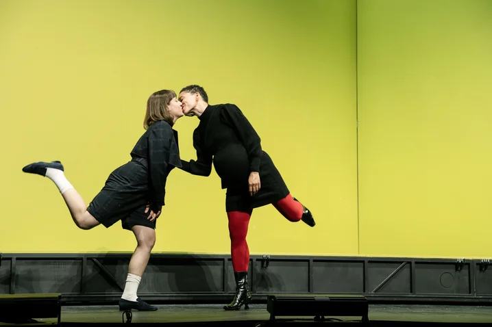 Two performers in black outfits dance in a modern, minimalist space with a bright green background.