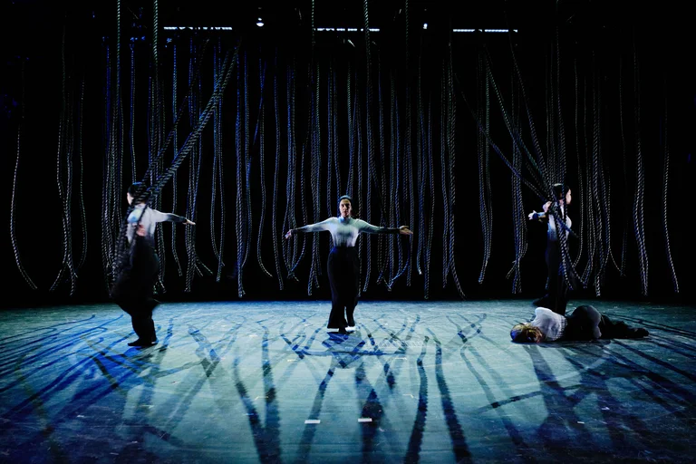 Tänzer auf einer dunklen Bühne mit langen, herabhängenden Stoffbahnen, die dramatische Schatten werfen. Eine Person liegt auf dem Boden.