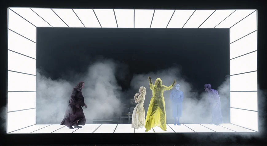 Performers in luminous costumes on a futuristic stage, surrounded by smoke and mist, against a dark backdrop.