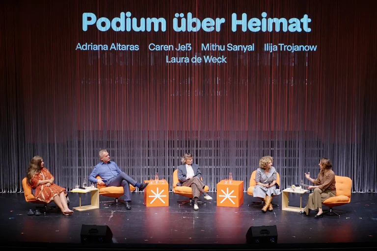 Eine Podiumsdiskussion mit fünf Personen auf einer Bühne. Der Hintergrund projiziert den Titel "Podium über Heimat".