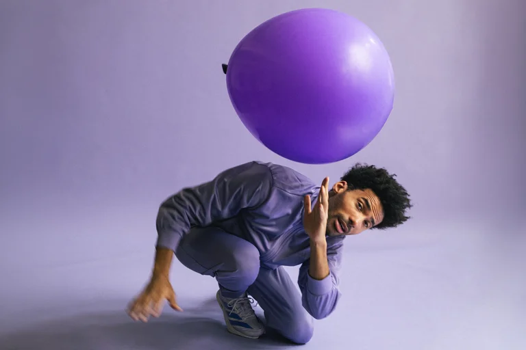 Ein Mann im grauen Anzug posiert in einem Studio mit einem großen lila Ballon über seinem Kopf.
