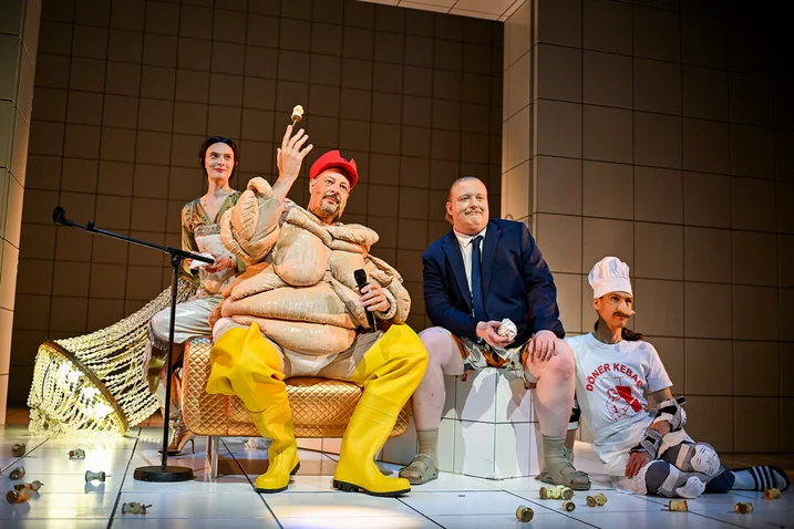 A theatrical scene with performers in elaborate costumes. One character wears a red hat and yellow boots.