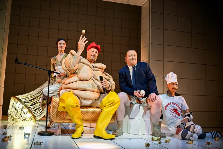 Eine theatralische Szene mit Darstellern in kunstvollen Kostümen. Eine Figur trägt einen roten Hut und gelbe Stiefel.