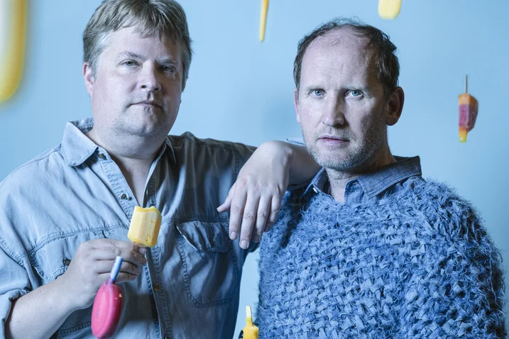 Two men stand against a blue background. They wear casual clothing, and colorful objects float around them.