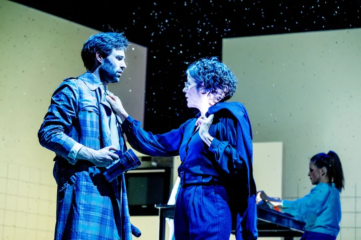 Two actors in a dramatic stage scene. A man in a coat, a woman gently touches him. Starry backdrop.