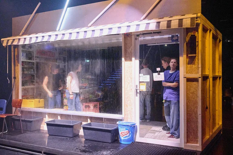 Eine kleine Bühne zeigt eine Ladenfront mit Menschen im Inneren, beleuchtet von neonartigen Lichtern. Eimer stehen draußen.