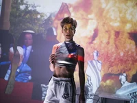 A performer stands before a projection of fire and people in traditional attire, wearing athletic clothing.