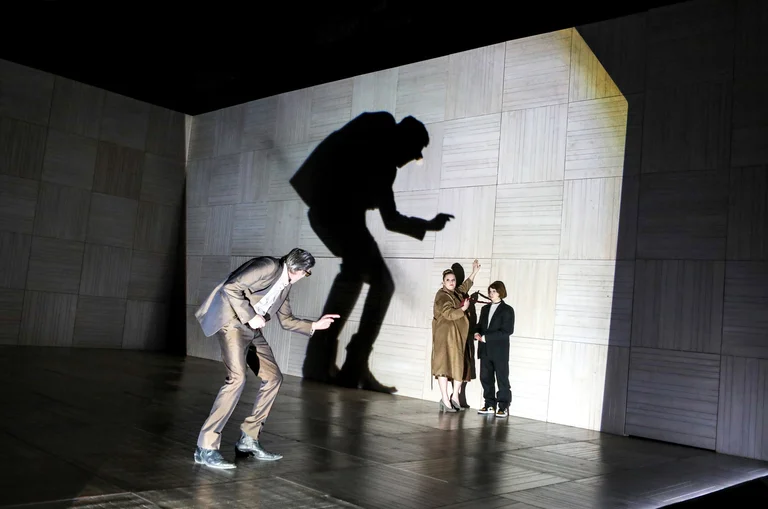 Eine Theaterszene mit drei Schauspielern. Eine übergroße Schattenfigur dominiert die Wand, während eine Frau und ein Kind zuschauen.
