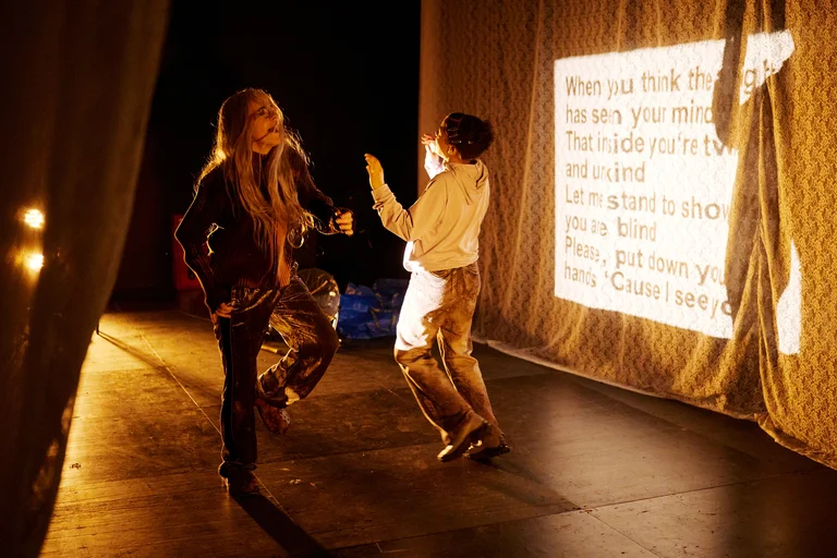 Zwei Personen tanzen leidenschaftlich auf einer Bühne, umgeben von warmem Licht. Im Hintergrund ist ein projizierter Text sichtbar.