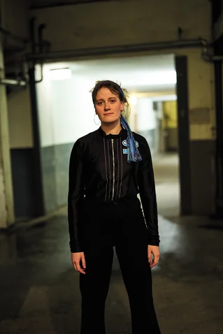 Eine Person steht in einem schlecht beleuchteten, industriell aussehenden Korridor. Sie trägt schwarze Kleidung mit blauen Akzenten.