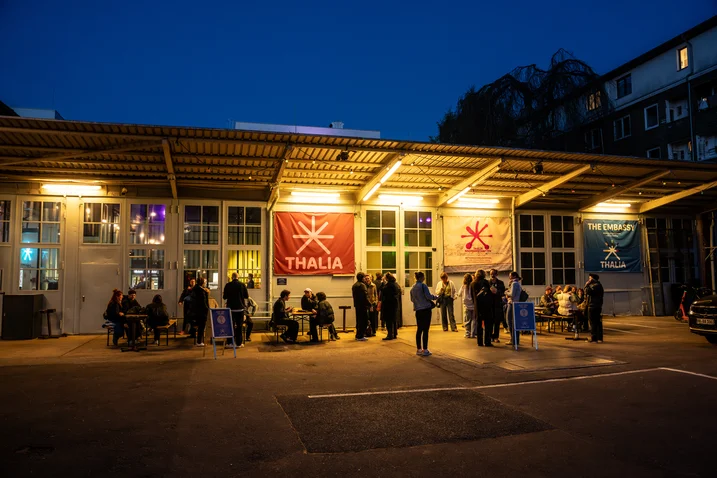 Abendliches Szene vor einem Gebäude mit beleuchteten Fenstern und Flaggen. Menschen stehen in Gruppen, die Atmosphäre wirkt lebhaft.
