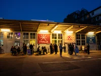 Abendliches Szene vor einem Gebäude mit beleuchteten Fenstern und Flaggen. Menschen stehen in Gruppen, die Atmosphäre wirkt lebhaft.