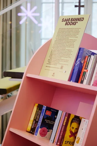 A bookshelf labeled "5,658 Banned Books" filled with various book titles. Bright, modern setting with artistic design elements.