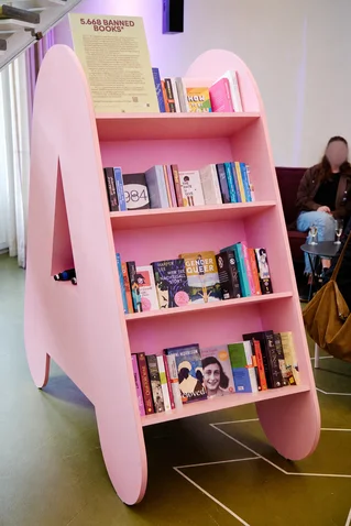 A pink bookshelf titled "365 Banned Books" filled with various books. Blurred people can be seen in the background.