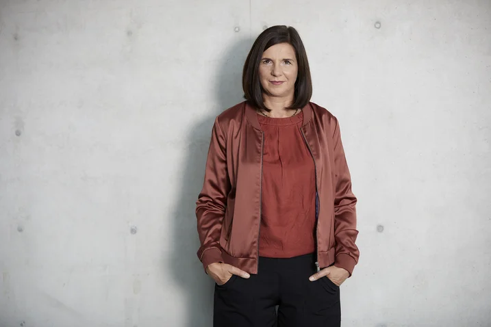 A woman in a red jacket poses against a gray background. She has shoulder-length dark hair, hands in pockets.
