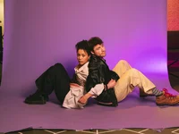Two people sit back-to-back on a purple backdrop. They wear stylish, casual clothing and have a serious expression.