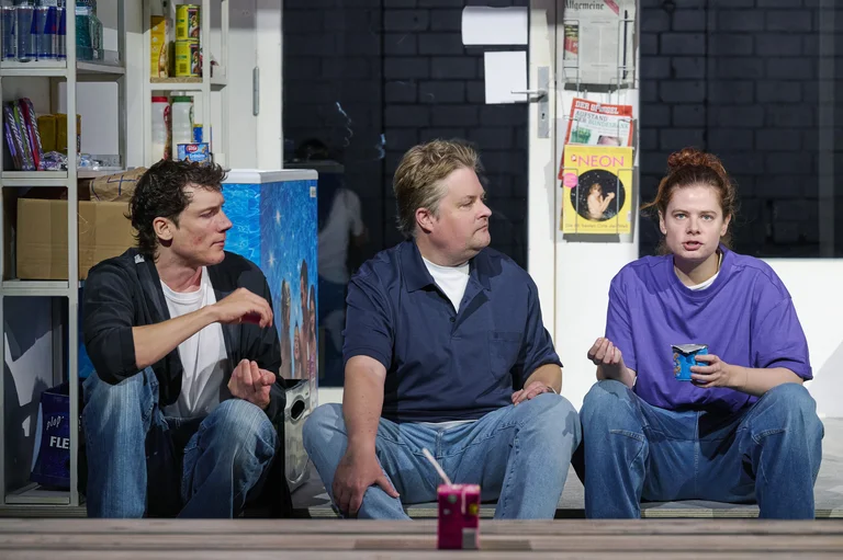 Drei Personen sitzen auf einer Bühne in einem Theaterstück. Sie diskutieren angeregt. Verschiedene Requisiten und ein Getränk sind sichtbar.