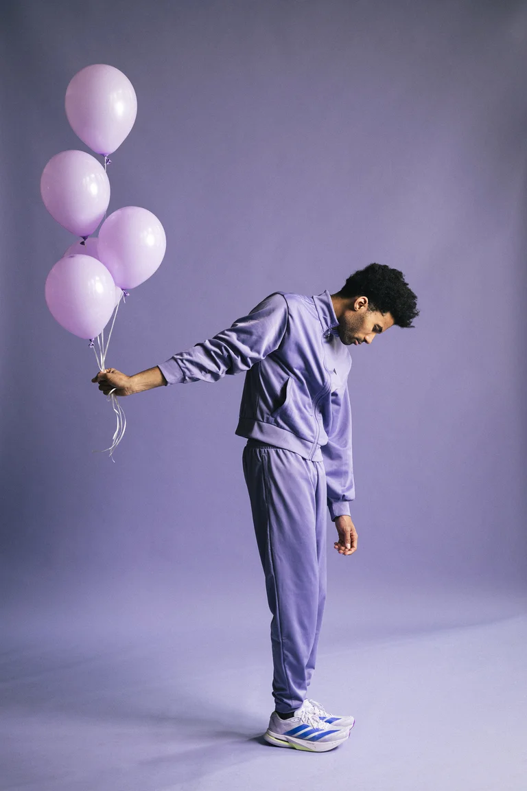 Ein Mann in einem lila Trainingsanzug hält vier rosa Luftballons in einem lila Raum. Er wirkt nachdenklich.