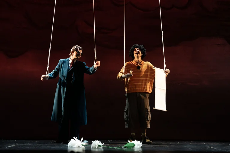 Zwei Darsteller auf der Bühne, als Marionetten mit Schnüren dargestellt, vor einem dunkelroten Hintergrund. Die Szene wirkt dramatisch.