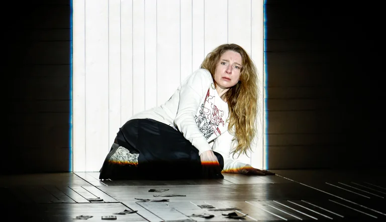 Eine Person mit langen Haaren sitzt auf dem Boden in einem dunklen Raum, beleuchtet von einem hellen Lichtstrahl.