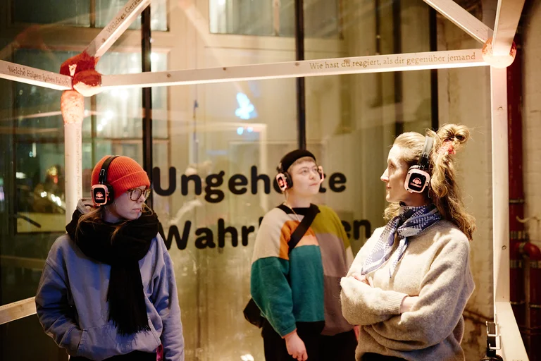 Three people wearing headphones stand in front of a glass box labeled "Ungehörte Wahrheiten", possibly communicating or listening.
