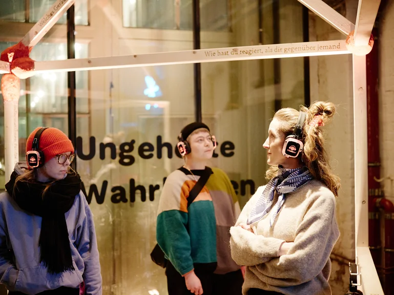 Three people wearing headphones stand in front of a glass box labeled "Ungehörte Wahrheiten", possibly communicating or listening.
