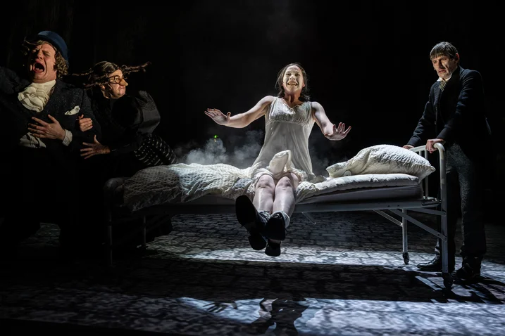 Theater scene with actors in dramatic pose on stage. One person lies on a bed, surrounded by three others.