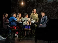 Six people sit at a round table on stage, with a dark backdrop and a single bulb illuminating the scene.