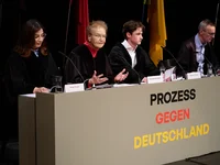Vier Personen sitzen an einem Tisch mit der Aufschrift "Prozess gegen Deutschland". Sie scheinen an einer Diskussion oder Debatte teilzunehmen.