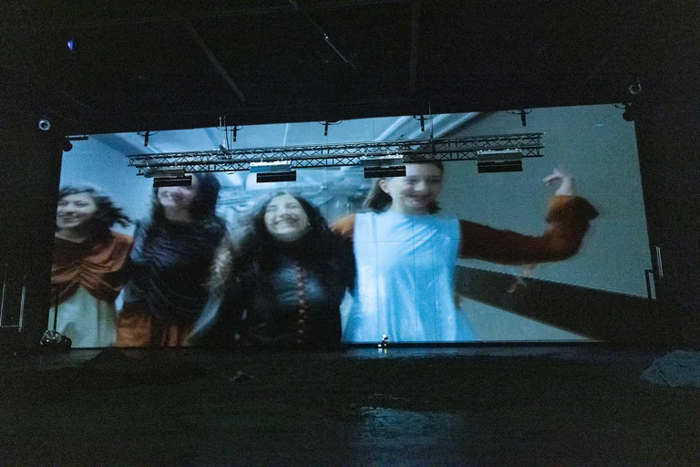 Eine Leinwand in einem dunklen Raum zeigt drei Menschen in Bewegung, die fröhliche Kleidung tragen, im Hintergrund technische Ausrüstung.