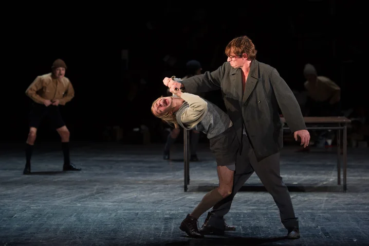 A dramatic dance scene on stage with two performers, one in a suit, another being swung. Background performances.