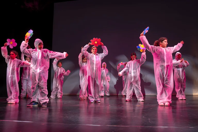 Kinder in rosa Kostümen tanzen auf einer Bühne. Sie halten bunte Accessoires und scheinen eine fröhliche Performance zu geben.
