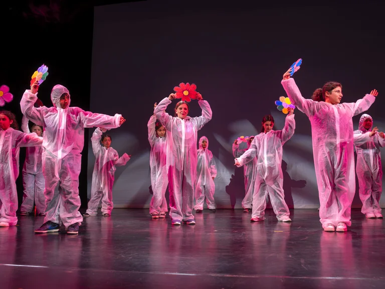 Kinder in rosa Kostümen tanzen auf einer Bühne. Sie halten bunte Accessoires und scheinen eine fröhliche Performance zu geben.