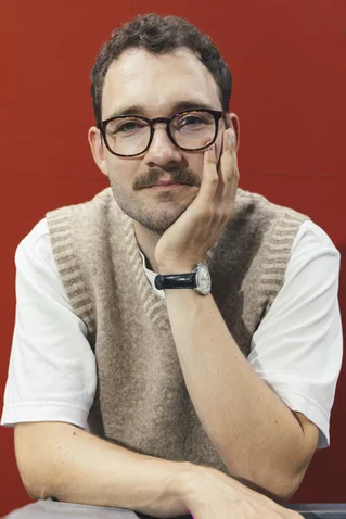 A person with glasses and a mustache sitting in front of a red background, resting their head on their hand.