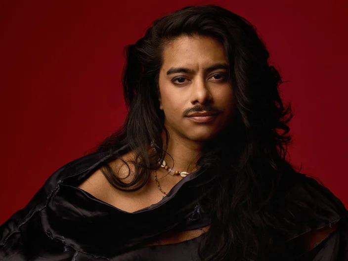 A person with long hair and a mustache stands against a red background, wearing dark, elegant attire.
