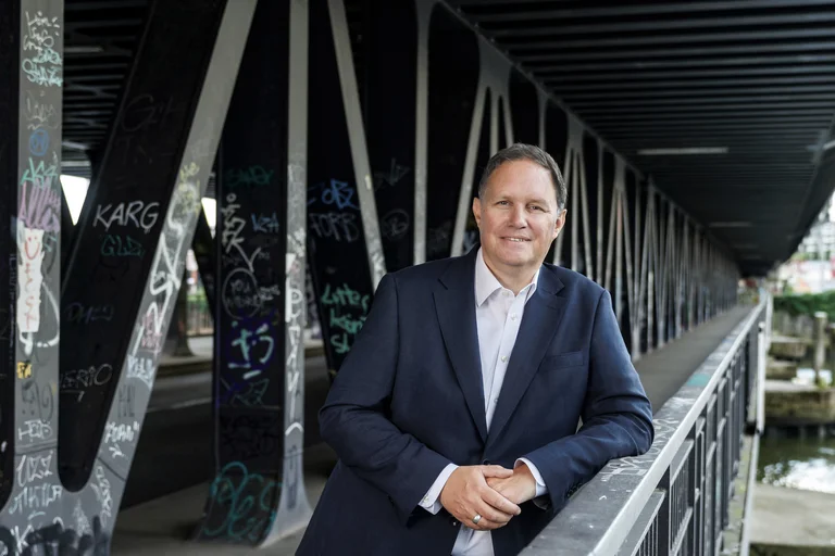Ein Mann im Anzug steht auf einer Brücke mit metallenen Strukturen, die von Graffiti bedeckt sind.
