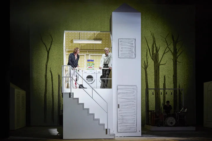 A stage with a staircase structure, two people, and a laundry room in a setting with painted trees.