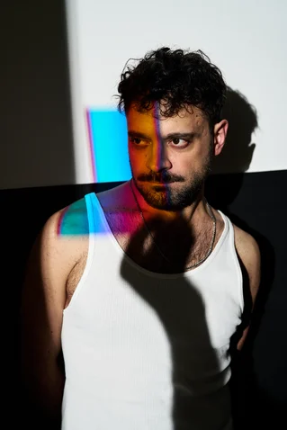 Ein Mann in einem weißen Tanktop steht im Schatten. Farbige Lichtprojektionen fallen auf sein Gesicht und den Hintergrund.
